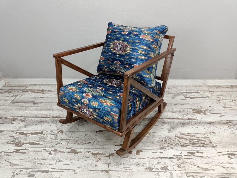 A wooden rocking chair with a blue and white patterned cushion, placed on a white wooden floor against a plain white wall.