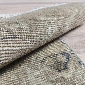May include: A close-up of a beige and brown woven rug with a subtle pattern. The rug is rolled up, showing the texture of the weave.