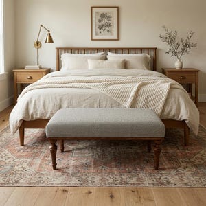May include: A bedroom scene featuring a wooden bed frame with a light-colored comforter and pillows. A gray upholstered bench sits at the foot of the bed. A patterned rug and two wooden nightstands complete the room's decor.