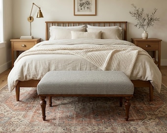 May include: A bedroom scene featuring a wooden bed frame with a light-colored comforter and pillows. A gray upholstered bench sits at the foot of the bed. A patterned rug and two wooden nightstands complete the room's decor.