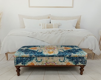 Op de afbeelding: Een blauwe en crèmekleurige opbergottoman met gouden accenten en houten poten. De ottoman staat voor een bed met witte beddengoed.