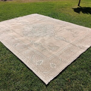 May include: A light beige rug with a detailed floral pattern in a lighter shade of beige. The rug has a fringed edge and is laid out on a green grassy surface.