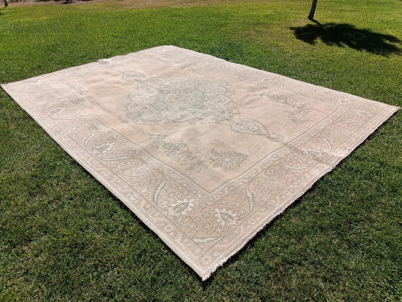 May include: A light beige rug with a detailed floral pattern in a lighter shade of beige. The rug has a fringed edge and is laid out on a green grassy surface.
