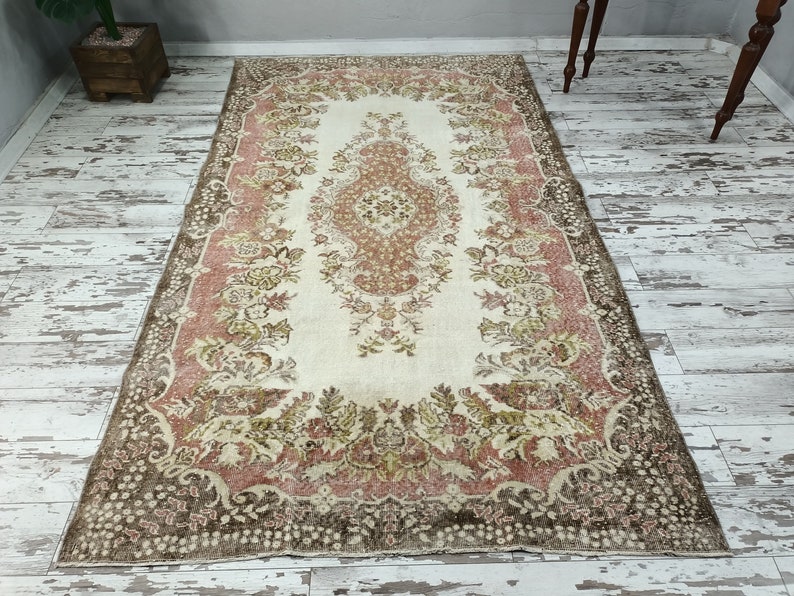 Op de afbeelding: Een vintage tapijt met een bloemenpatroon in roze, bruin en cr&egrave;me tinten. Het tapijt heeft een centraal medaillon en een rand met een herhalend ontwerp.
