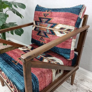 K&ouml;nnte beinhalten: Holzsessel mit gemustertem Kissen und R&uuml;ckenlehne. Das Kissen und die R&uuml;ckenlehne weisen ein Design mit roten, blauen und wei&szlig;en geometrischen Formen auf. Der Stuhl hat ein dunkles Holzgestell und steht vor einer Pflanze.