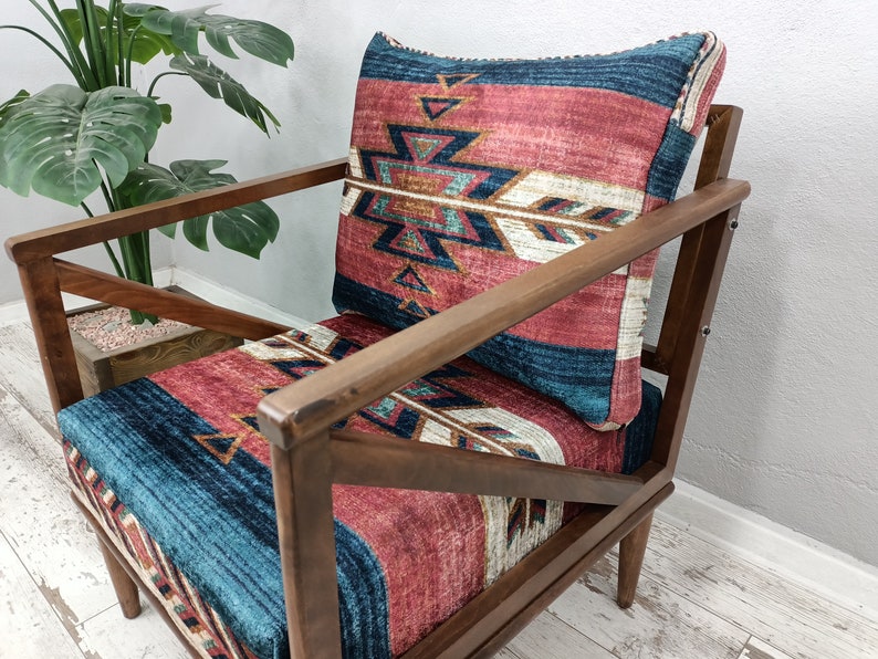 K&ouml;nnte beinhalten: Holzsessel mit gemustertem Kissen und R&uuml;ckenlehne. Das Kissen und die R&uuml;ckenlehne weisen ein Design mit roten, blauen und wei&szlig;en geometrischen Formen auf. Der Stuhl hat ein dunkles Holzgestell und steht vor einer Pflanze.