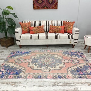 Pode incluir: Um tapete rosa e azul com um padr&atilde;o floral. O tapete est&aacute; colocado sobre um piso de madeira branca em frente a um sof&aacute; branco com almofadas listradas.