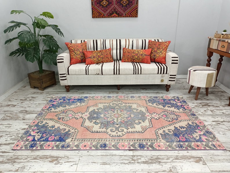 Pode incluir: Um tapete rosa e azul com um padr&atilde;o floral. O tapete est&aacute; colocado sobre um piso de madeira branca em frente a um sof&aacute; branco com almofadas listradas.
