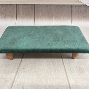 Puede incluir: Un reposapiés rectangular de color verde oscuro con una parte superior de tela suave y texturizada y cuatro patas de madera.