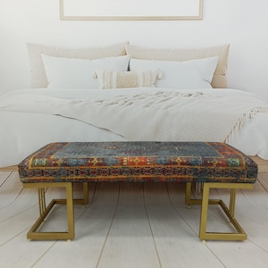 May include: A rectangular upholstered bench with a patterned top in shades of blue, red, and gold. The bench has a gold metal frame and is placed at the foot of a bed with white bedding. The room has a neutral color palette.