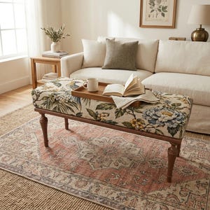 May include: An upholstered rectangular bench with a cream-colored fabric featuring a botanical print of blue and yellow flowers and green leaves. The bench has four dark brown wooden legs and supports a wooden tray with a white mug and an open book.