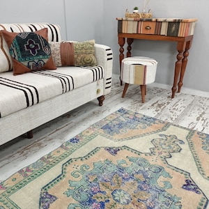 K&ouml;nnte beinhalten: Ein wei&szlig;-braunes gestreiftes Sofa mit einem gr&uuml;n-braunen gemusterten Kissen, einem braun-wei&szlig;en gemusterten Kissen und einem beige-wei&szlig;en gemusterten Kissen. Das Sofa steht auf einem hellen Holzboden mit einem hellblau-rosa gemusterten Teppich davor. Im Hintergrund steht ein kleiner Holztisch mit einem wei&szlig;-braunen gemusterten Hocker.