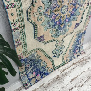 K&ouml;nnte beinhalten: Ein Vintage-T&uuml;rkischer Teppich mit cremefarbenem Hintergrund und einem blauen, gr&uuml;nen und rosafarbenen Blumenmuster. Der Teppich hat einen abgenutzten Look und ist perfekt, um Ihrem Zuhause einen Hauch von Boho-Stil zu verleihen.