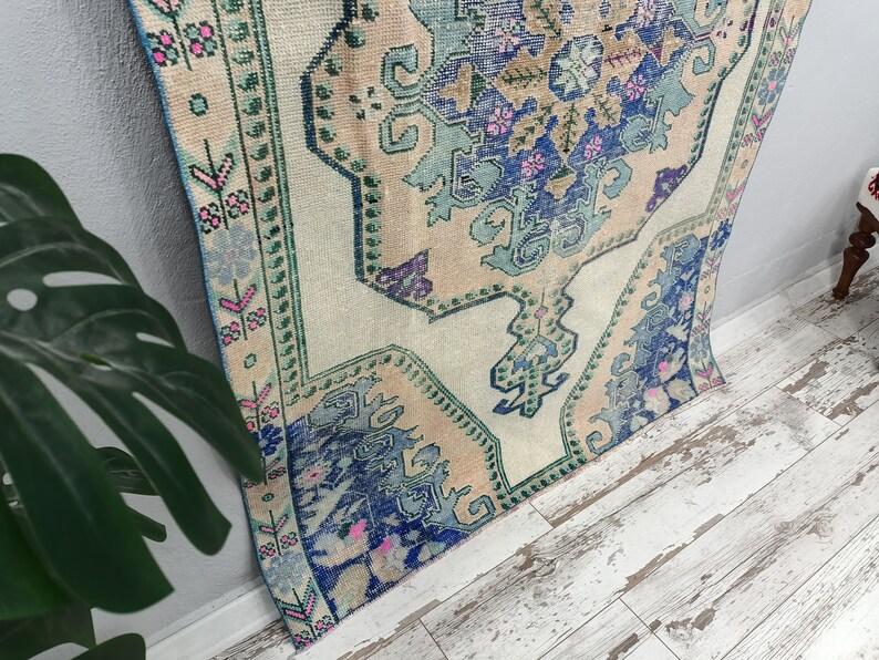 K&ouml;nnte beinhalten: Ein Vintage-T&uuml;rkischer Teppich mit cremefarbenem Hintergrund und einem blauen, gr&uuml;nen und rosafarbenen Blumenmuster. Der Teppich hat einen abgenutzten Look und ist perfekt, um Ihrem Zuhause einen Hauch von Boho-Stil zu verleihen.