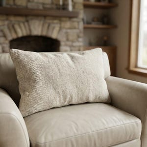 May include: A rectangular beige textured pillow rests on a light beige armchair. The pillow has a woven appearance and is slightly rumpled. The armchair is made of a smooth material, and the background includes a fireplace and a window.