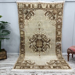 May include: A vintage rug with a cream background and a brown floral pattern. The rug has a fringe border and is hanging on a wall.