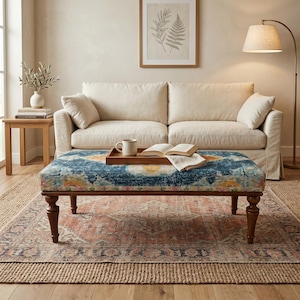 May include: A living room scene featuring a cream-colored sofa, a patterned ottoman, and a vintage-style rug. The ottoman has a blue, yellow, and orange design. A wooden tray with a mug and book sits on the ottoman. A floor lamp provides soft lighting.