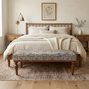 Op de afbeelding: Een slaapkamer met een houten bed, neutrale beddengoed en een bank met patroon. De kamer omvat een vintage tapijt, een ingelijste botanische print en een messing wandlamp. De sfeer is warm en uitnodigend.