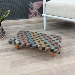 May include: A rectangular footstool with a woven, checkered pattern in shades of brown, blue, yellow, and black. The footstool has four short, rounded wooden legs. The item is placed on a light wood floor.