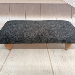 May include: A black fabric footstool with wooden legs. The fabric has a subtle pattern.
