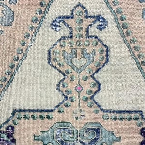 K&ouml;nnte beinhalten: Nahaufnahme eines hellrosa und wei&szlig;en Teppichs mit einem blauen und gr&uuml;nen geometrischen Muster. Das Muster zeigt ein zentrales Design mit einer Herzform und einer Reihe von Punkten.