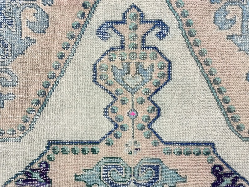 K&ouml;nnte beinhalten: Nahaufnahme eines hellrosa und wei&szlig;en Teppichs mit einem blauen und gr&uuml;nen geometrischen Muster. Das Muster zeigt ein zentrales Design mit einer Herzform und einer Reihe von Punkten.