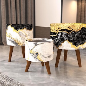 May include: Three cylindrical stools with tapered wooden legs, featuring a patterned exterior of black, white, and gold marble-like textures. The stools vary in height and diameter, each displaying a distinct arrangement of the marbled design.