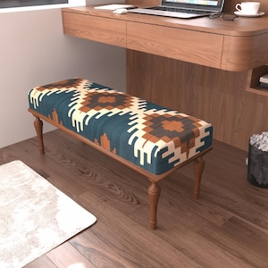 May include: A rectangular upholstered bench featuring a geometric pattern in shades of teal, brown, and cream. The seat is supported by four turned wooden legs in a medium brown finish, providing a stable base for this piece of furniture.