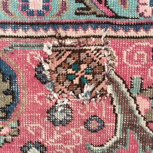 May include: Close-up of a pink and blue patterned rug with a small tear in the center. The tear is a rectangular shape and is surrounded by frayed edges.