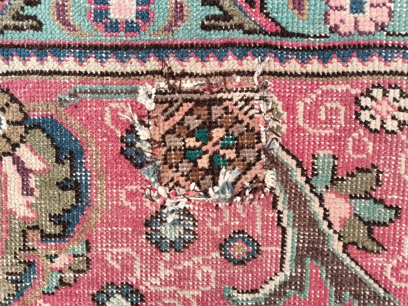 May include: Close-up of a pink and blue patterned rug with a small tear in the center. The tear is a rectangular shape and is surrounded by frayed edges.