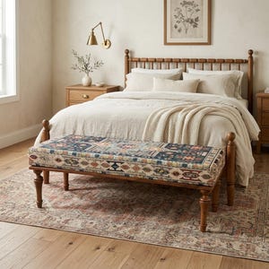 May include: A bedroom scene featuring a wooden bench with a patterned cushion at the foot of a bed. The bench has four turned legs and a rectangular cushion with a dark blue, beige, and rust-colored design. The bed has a light-colored comforter and pillows.