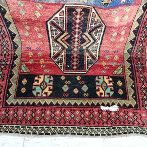 May include: A red, black, and green patterned rug with a geometric design. The rug has a fringe border.