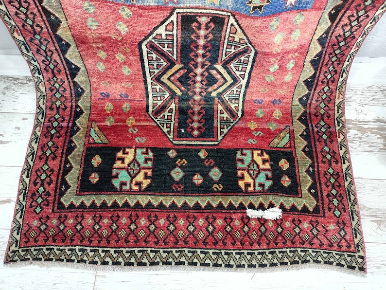 May include: A red, black, and green patterned rug with a geometric design. The rug has a fringe border.