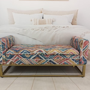 May include: A patterned bench with a gold metal frame. The bench features a colorful geometric design in shades of blue, red, yellow, and brown. The bench is placed in front of a bed with white bedding.
