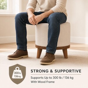 A man sitting on a white ottoman with a wood frame, wearing brown shoes and blue jeans.