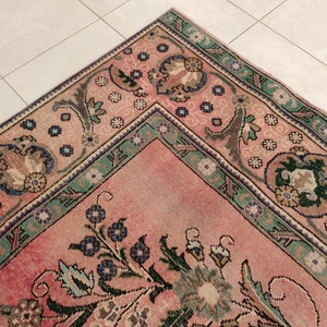 May include: A vintage rug with a pink background and intricate floral patterns in shades of green, blue, and brown. The rug has a border with a repeating design of flowers and leaves.