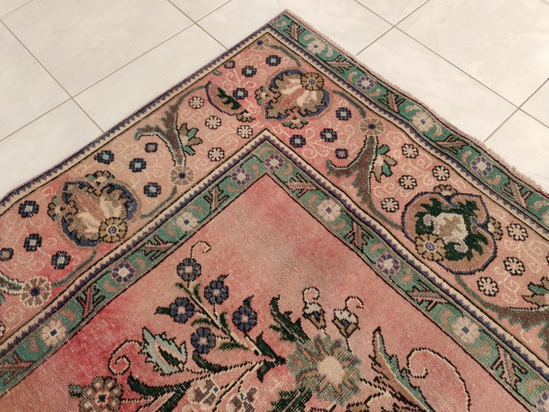 May include: A vintage rug with a pink background and intricate floral patterns in shades of green, blue, and brown. The rug has a border with a repeating design of flowers and leaves.
