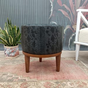 May include: A black faux leather ottoman with a snake skin pattern and wooden legs. The ottoman is sitting on a pink and beige rug.