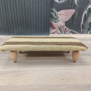 May include: A rectangular footstool with a beige and brown striped pattern. The stool has four short, rounded wooden legs. The overall design is simple and rustic, suitable for a living room or bedroom.