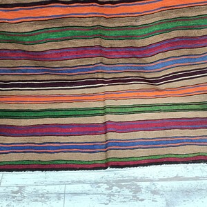 May include: A striped woven rug with a variety of colors, including brown, orange, green, blue, and red. The rug has a fringed edge and is made of natural fibers.