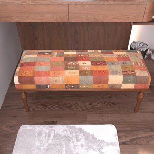 a wooden bench with a colorful, patchwork-like cushion on it, placed on a wooden floor. There is a small pile of white items next to the bench, and a white rug is visible in the foreground.