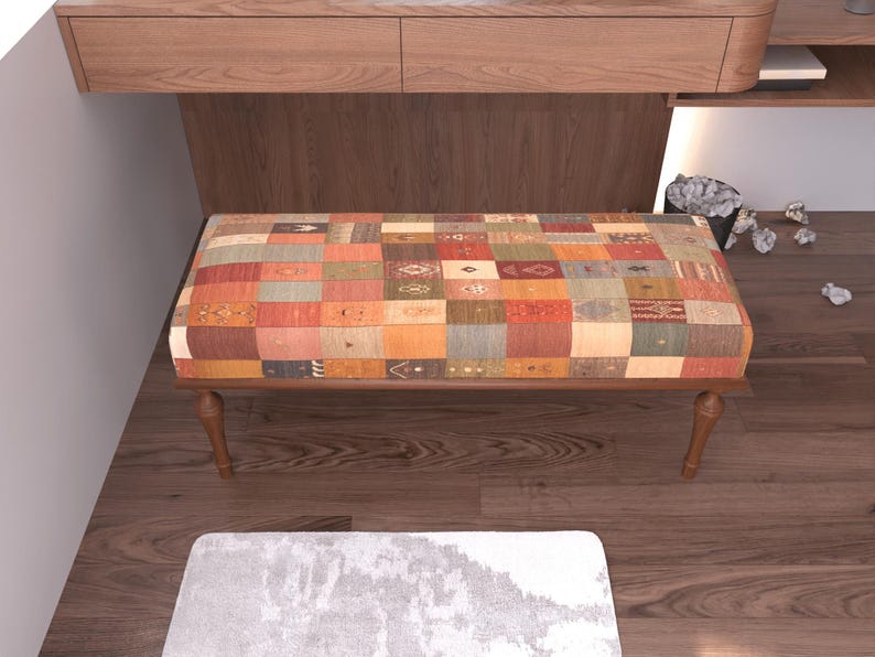 a wooden bench with a colorful, patchwork-like cushion on it, placed on a wooden floor. There is a small pile of white items next to the bench, and a white rug is visible in the foreground.