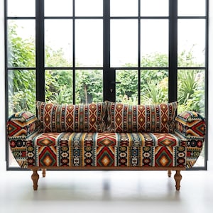 May include: A sofa with a wooden frame and turned legs. The upholstery has a geometric pattern in red, yellow, blue, and black. Two matching throw pillows are on the sofa. The sofa is in front of a large window.