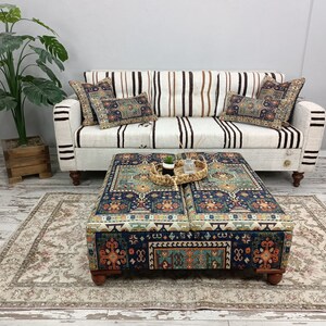 May include: A white striped sofa with two patterned throw pillows sits in front of a white wall. A patterned ottoman with a woven tray on top sits in front of the sofa. A beige rug with a floral pattern is on the floor.