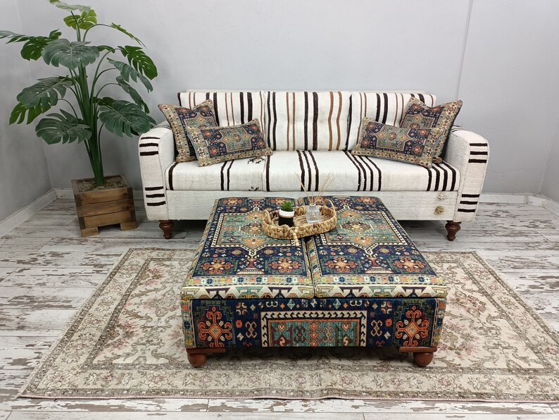 May include: A white striped sofa with two patterned throw pillows sits in front of a white wall. A patterned ottoman with a woven tray on top sits in front of the sofa. A beige rug with a floral pattern is on the floor.