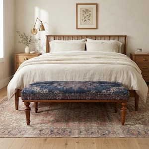 a cozy bedroom with a large bed, a wooden headboard, and a wooden nightstand. The bed is covered in a white comforter and has a blue patterned bench in front of it. The room has a wooden floor and a framed picture on the wall above the bed.