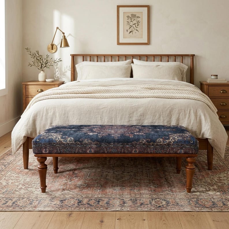 a cozy bedroom with a large bed, a wooden headboard, and a wooden nightstand. The bed is covered in a white comforter and has a blue patterned bench in front of it. The room has a wooden floor and a framed picture on the wall above the bed.