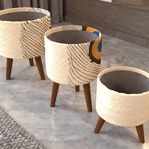 May include: Three cylindrical planters with three wooden legs each. The exterior features a cream-colored textured surface with black line patterns and a partial peacock feather design in blue, gold, and brown. The interior is lined with gray fabric.