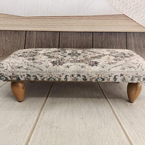 A decorative ottoman with a patterned fabric and wooden legs, placed on a wooden floor against a wooden background.