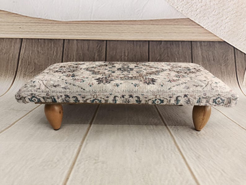 A decorative ottoman with a patterned fabric and wooden legs, placed on a wooden floor against a wooden background.